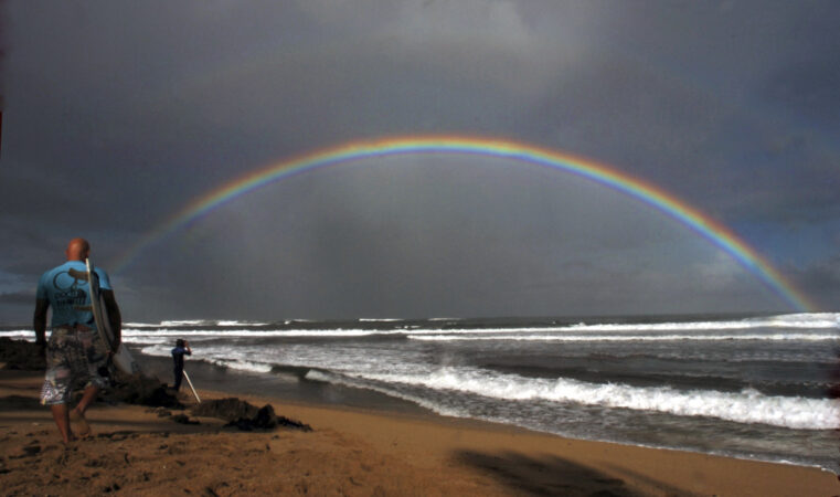Surfers make their way to the water while a large rainbow crosses the ocean in Hawaii.